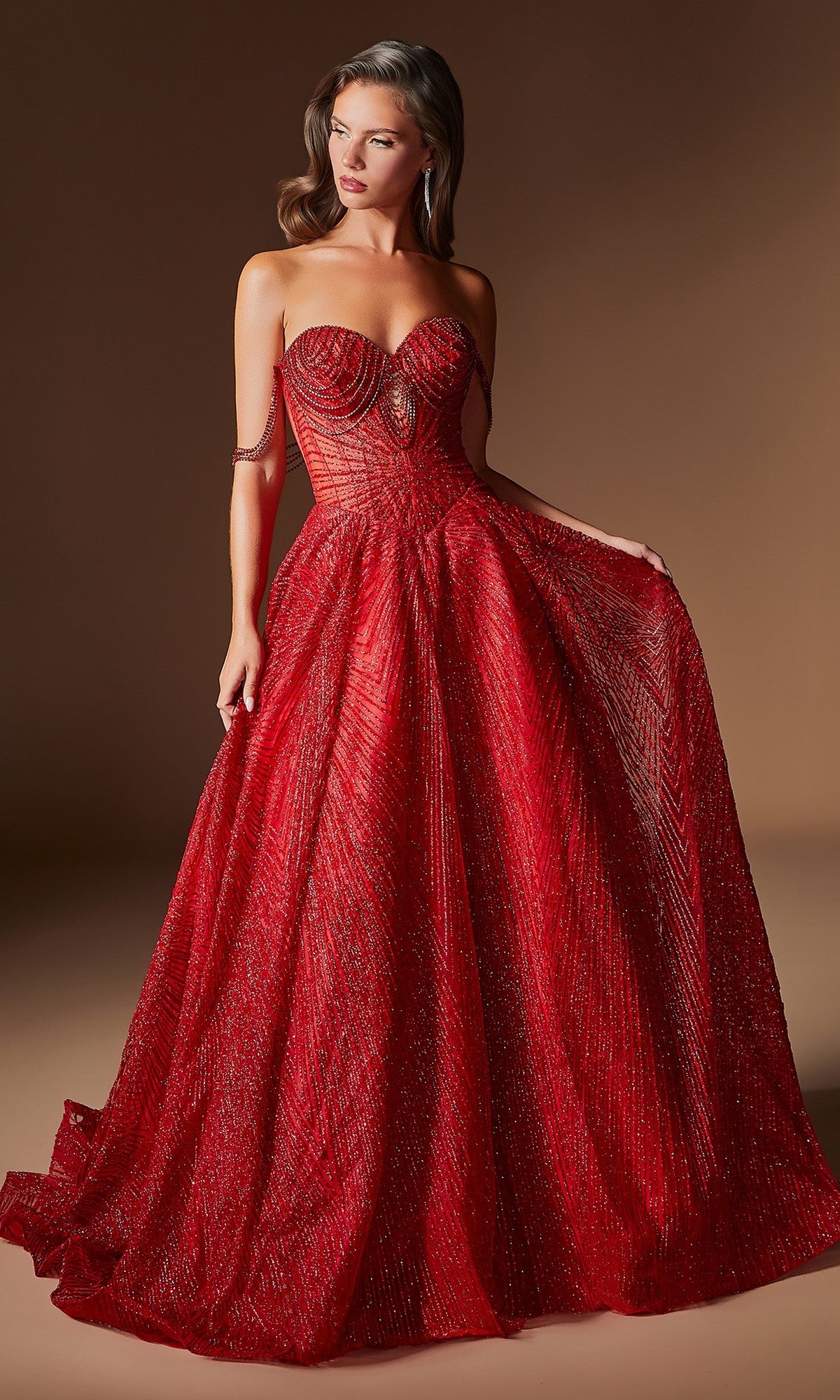 A woman wearing the Ladivine J915 long red prom dress, which features a strapless neckline, textured fabric with intricate patterns and crystal beadwork, stands against a brown background while holding one side of the gown.